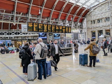 A busy train station with people gathered around a digital departure board, checking schedules. Travelers with luggage are walking through the hall, and some are sitting on benches in the background. The architectural details include a high, arched ceiling with industrial design elements.