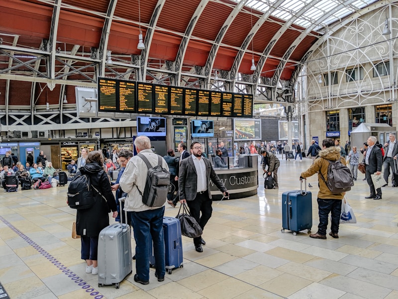 A busy train station with people gathered around a digital departure board, checking schedules. Travelers with luggage are walking through the hall, and some are sitting on benches in the background. The architectural details include a high, arched ceiling with industrial design elements.
