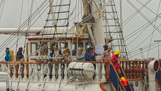 People are standing on the deck of a large sailing ship, surrounded by numerous ropes and rigging. The ship has a classic wood and white design with lifeboats attached. Some individuals are wearing jackets and hats, suggesting cool weather conditions.