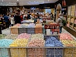 A bustling candy store with a colorful display of individually wrapped chocolates arranged in open bins. Shoppers are selecting candies from the wide variety, while others browse the shelves filled with sweet treats. The store has a warm and inviting atmosphere.