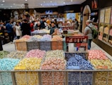 A bustling candy store with a colorful display of individually wrapped chocolates arranged in open bins. Shoppers are selecting candies from the wide variety, while others browse the shelves filled with sweet treats. The store has a warm and inviting atmosphere.