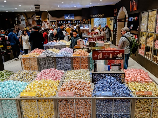A bustling candy store with a colorful display of individually wrapped chocolates arranged in open bins. Shoppers are selecting candies from the wide variety, while others browse the shelves filled with sweet treats. The store has a warm and inviting atmosphere.