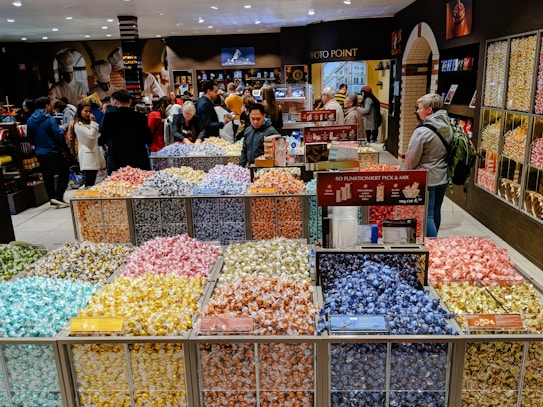 A bustling candy store with a colorful display of individually wrapped chocolates arranged in open bins. Shoppers are selecting candies from the wide variety, while others browse the shelves filled with sweet treats. The store has a warm and inviting atmosphere.