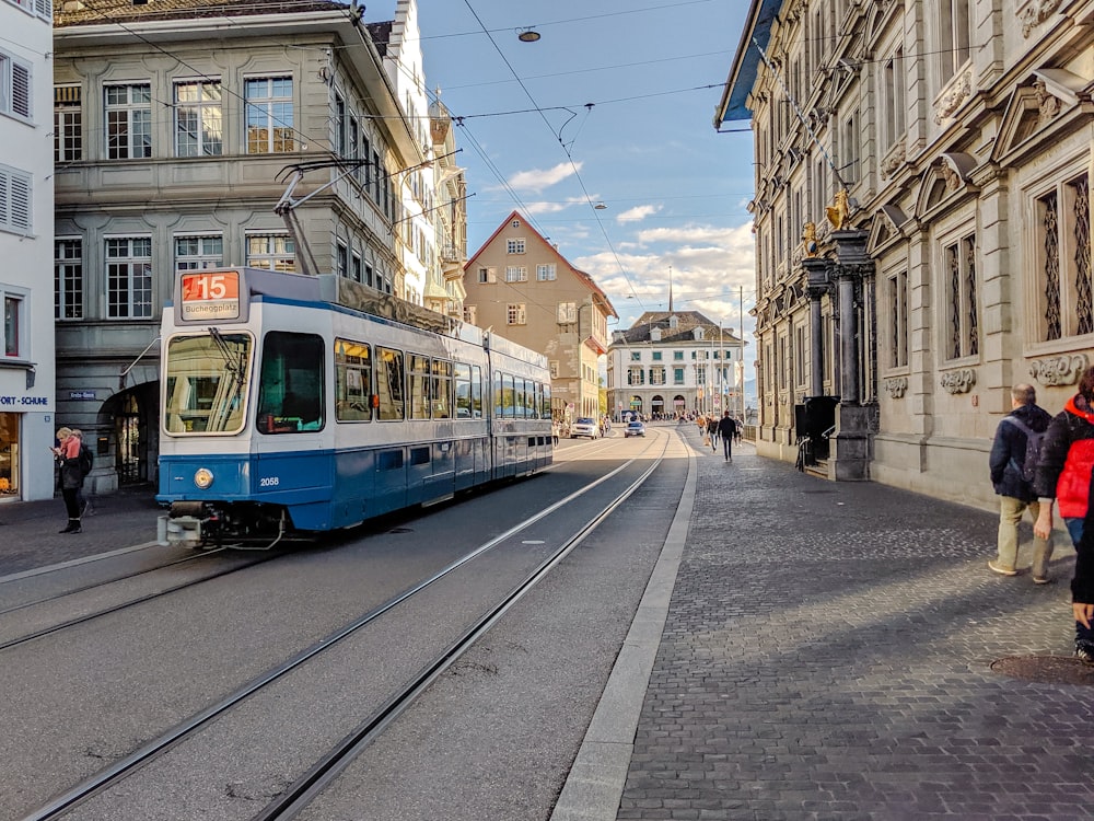 a blue and white train traveling down a street next to tall buildings