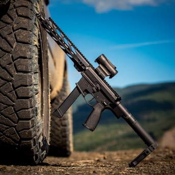 A black rifle is leaning against an off-road vehicle tire. The setting appears to be outdoors, with a clear blue sky and distant mountains in the background.
