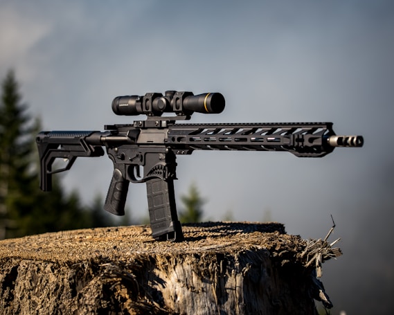 A modern rifle with a scope is positioned on top of a large tree stump. The background features an out-of-focus view of trees under a clear sky, giving the image an outdoor setting.
