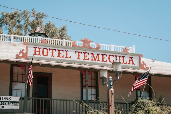 An old-style building with a wooden facade and a sign that reads 'Hotel Temecula' in large red letters. Two American flags are displayed, one on each side of the balcony railing. The structure includes vintage-style street lamps and greenery in the background.