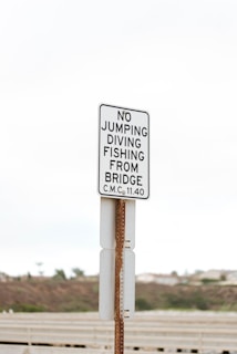 A white and black sign is posted on a metal pole. The sign contains instructions prohibiting activities such as jumping, diving, and fishing from a bridge. The background shows an overcast sky and blurred foliage, suggesting an outdoor location near a bridge.