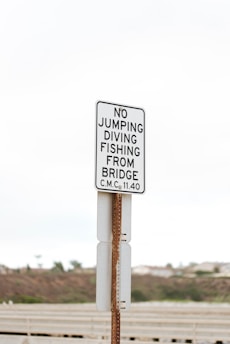 A white and black sign is posted on a metal pole. The sign contains instructions prohibiting activities such as jumping, diving, and fishing from a bridge. The background shows an overcast sky and blurred foliage, suggesting an outdoor location near a bridge.