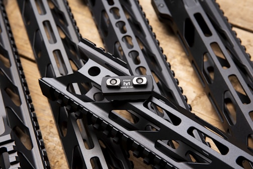 Several sleek, black metallic handguards featuring cut-out designs are arranged in a diagonal pattern on a wooden surface. A small metal component labeled 'STRIKE M MADE IN USA' is prominently attached to one of the handguards.