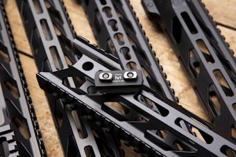 Several sleek, black metallic handguards featuring cut-out designs are arranged in a diagonal pattern on a wooden surface. A small metal component labeled 'STRIKE M MADE IN USA' is prominently attached to one of the handguards.