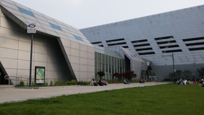 A modern architectural structure with angular and geometric shapes dominates the scene, featuring a facade with small windows. People are sitting and interacting on a paved area surrounded by a neatly maintained lawn, creating a contrast between the building's hard lines and the soft grass. A signpost with a logo and some text in another language is visible, along with some landscaped vegetation.