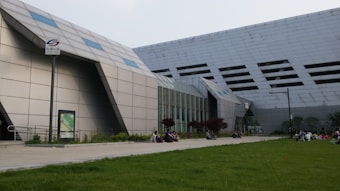 A modern architectural structure with angular and geometric shapes dominates the scene, featuring a facade with small windows. People are sitting and interacting on a paved area surrounded by a neatly maintained lawn, creating a contrast between the building's hard lines and the soft grass. A signpost with a logo and some text in another language is visible, along with some landscaped vegetation.