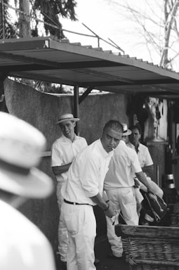 A group of men dressed in white clothing and hats appear to be engaged in an outdoor activity under a shaded area. One man seems to be bending over a wicker basket, suggesting involvement in some form of physical task. The atmosphere appears candid and informal with some men smiling.