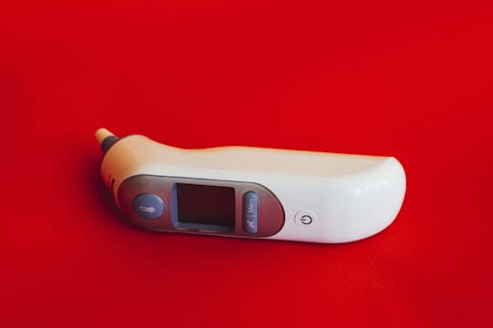 A digital thermometer lies on a bright red surface. The thermometer features a sleek, ergonomic design with a white body, a small display screen, and several buttons.