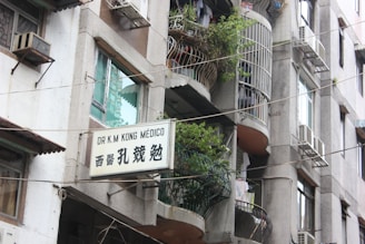 A multi-story building with balconies adorned with potted plants and laundry. Several air conditioning units are installed on the walls. A sign in both English and Chinese characters is displayed, indicating the presence of a medical practice.