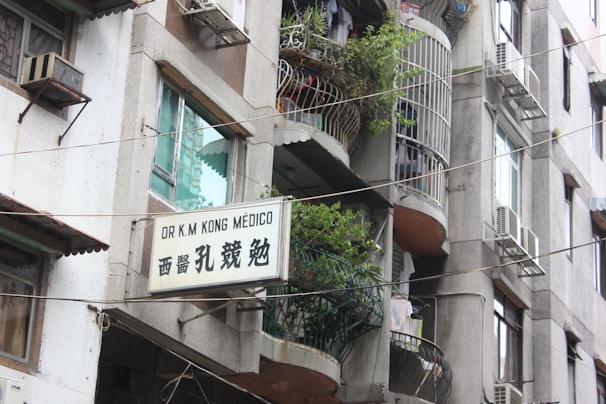 A multi-story building with balconies adorned with potted plants and laundry. Several air conditioning units are installed on the walls. A sign in both English and Chinese characters is displayed, indicating the presence of a medical practice.