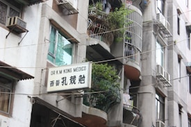 A multi-story building with balconies adorned with potted plants and laundry. Several air conditioning units are installed on the walls. A sign in both English and Chinese characters is displayed, indicating the presence of a medical practice.