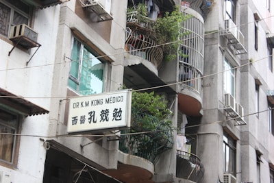 A multi-story building with balconies adorned with potted plants and laundry. Several air conditioning units are installed on the walls. A sign in both English and Chinese characters is displayed, indicating the presence of a medical practice.