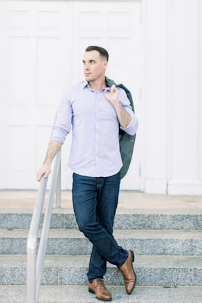 A man stands confidently on stone steps, leaning casually against a railing. He is dressed in a light blue button-up shirt and dark jeans, with a suit jacket slung over his shoulder. His posture is relaxed, suggesting a poised and self-assured demeanor.