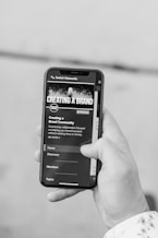 A hand holding a smartphone displaying a website or app interface related to creating a brand. The screen shows options for Home, Discovery, Members, and Topics.