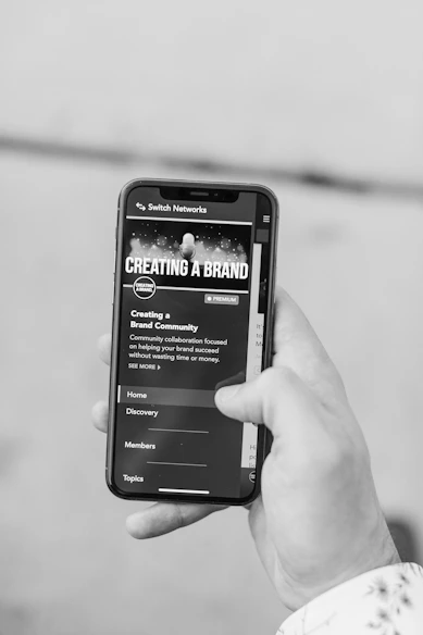A hand holding a smartphone displaying a website or app interface related to creating a brand. The screen shows options for Home, Discovery, Members, and Topics.