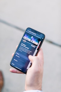 A hand holding a smartphone displaying a webpage titled 'Creating a Brand' with navigation options like Home, Discovery, Members, and Topics. The background is out of focus, providing a neutral setting.