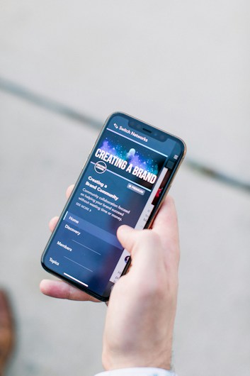 A hand holding a smartphone displaying a webpage titled 'Creating a Brand' with navigation options like Home, Discovery, Members, and Topics. The background is out of focus, providing a neutral setting.
