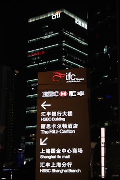 A nighttime cityscape features tall buildings, prominently displaying the logos of international banks such as HSBC and Citi, with bright lights and text guiding visitors to various locations including the HSBC Building and the Ritz-Carlton.