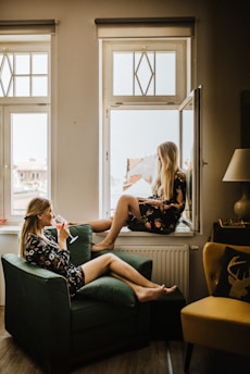Two women are relaxing in a cozy interior space. One is sitting on a green armchair, sipping a drink, and the other is perched on a windowsill with a book in her lap. They are both wearing floral dresses and there is a soft, warm light coming in from the windows, adding to the tranquil atmosphere.