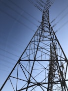 A tall metal transmission tower stretches into the sky, supported by a series of intricate steel beams. Multiple power lines extend from the tower, crisscrossing against the backdrop of a clear blue sky.