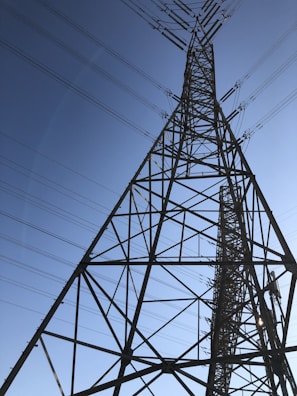 A tall metal transmission tower stretches into the sky, supported by a series of intricate steel beams. Multiple power lines extend from the tower, crisscrossing against the backdrop of a clear blue sky.