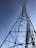 A tall metal transmission tower stretches into the sky, supported by a series of intricate steel beams. Multiple power lines extend from the tower, crisscrossing against the backdrop of a clear blue sky.