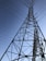 A tall metal transmission tower stretches into the sky, supported by a series of intricate steel beams. Multiple power lines extend from the tower, crisscrossing against the backdrop of a clear blue sky.