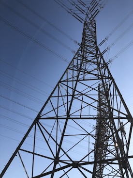 A tall metal transmission tower stretches into the sky, supported by a series of intricate steel beams. Multiple power lines extend from the tower, crisscrossing against the backdrop of a clear blue sky.