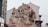 A detailed stone sculpture featuring multiple figures in traditional attire. The figures are intricately carved, positioned in dynamic and expressive poses, suggesting a narrative or historical scene. It is set in an urban environment, with signs and buildings visible in the background.