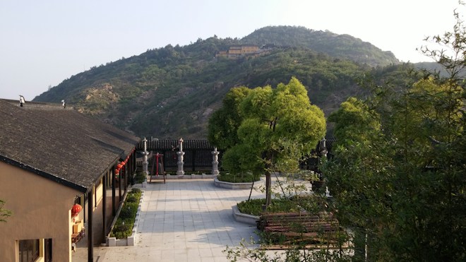 A peaceful courtyard is surrounded by lush green trees, with a traditional building on the left adorned with red lanterns. In the distance, a green hill with a few visible structures rises under a clear sky.