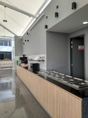 A modern kitchen space with a long countertop made of dark granite. The cabinetry below is made of light wood. Stainless steel food trays with lids are neatly arranged on the counter. On the back wall, there is a coffee machine, a microwave, and stacked dishes. The walls are painted gray, and the ceiling features several simple black pendant lights. Natural light enters through large windows on the left.