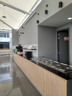 A modern kitchen space with a long countertop made of dark granite. The cabinetry below is made of light wood. Stainless steel food trays with lids are neatly arranged on the counter. On the back wall, there is a coffee machine, a microwave, and stacked dishes. The walls are painted gray, and the ceiling features several simple black pendant lights. Natural light enters through large windows on the left.