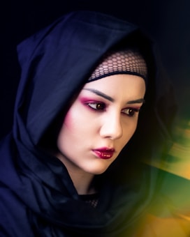 A person with distinctive makeup, featuring bold pink eyeshadow and dark lipstick, is wearing a black hooded garment and a netted headpiece. The lighting creates dramatic shadows on their face, and there is a soft blur of green and yellow hues on the right side.