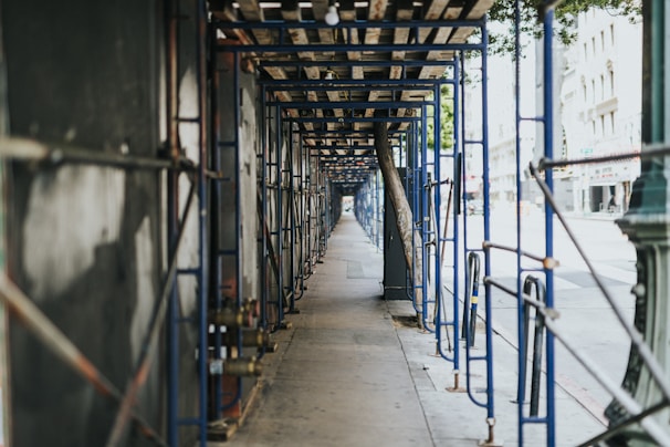 A long, symmetrical corridor of blue scaffolding lines a sidewalk, casting shadows along the ground. The scaffolding is designed to support ongoing construction or maintenance on an adjacent building. There is a clear perspective view leading down the length of the temporary structure, with trees and the facade of a city street visible in the background.