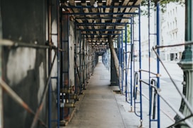 A long, symmetrical corridor of blue scaffolding lines a sidewalk, casting shadows along the ground. The scaffolding is designed to support ongoing construction or maintenance on an adjacent building. There is a clear perspective view leading down the length of the temporary structure, with trees and the facade of a city street visible in the background.
