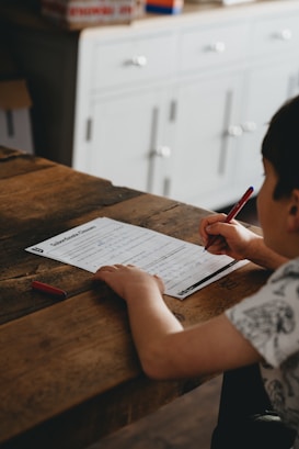 A child is sitting at a wooden table, concentrating on a worksheet with a pen in hand. The setting appears to be a room with a light-colored cabinet in the background, and there is a small red eraser on the table.