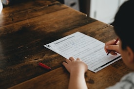 A person is writing on a worksheet titled 'Subordinate Clauses' placed on a wooden table. The individual holds a red pen and is filling in answers. A red pen cap lies nearby on the table.