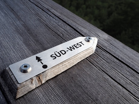 A metallic compass plaque mounted on a wooden surface, indicating the direction 'Süd-West' with an arrow and circle symbol.