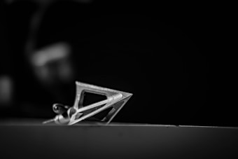 A metallic three-bladed arrowhead lies in sharp focus against a blurred dark background, showcasing its intricate and sharp design.