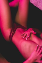 A figure lies back with legs bent at the knees, illuminated by a soft pink light. Hands rest gently on the torso, and the scene is intimate and artistic, with a focus on form and lighting.