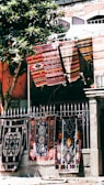 Colorful, patterned textiles hang over a metal fence in an outdoor setting. Several rugs are displayed, showcasing intricate designs and vibrant colors. A nearby tree with green leaves provides partial shade, and a stone building with textured walls stands in the background.