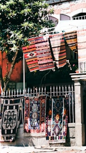 Colorful, patterned textiles hang over a metal fence in an outdoor setting. Several rugs are displayed, showcasing intricate designs and vibrant colors. A nearby tree with green leaves provides partial shade, and a stone building with textured walls stands in the background.