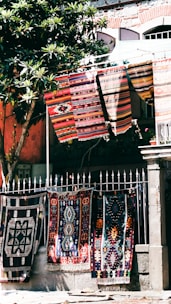 Colorful, patterned textiles hang over a metal fence in an outdoor setting. Several rugs are displayed, showcasing intricate designs and vibrant colors. A nearby tree with green leaves provides partial shade, and a stone building with textured walls stands in the background.